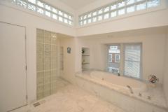 Master bathroom, marble tiled, high glass block windows allow for natural light, double vanity, full length mirror, jetted tub with hand sprayer, built-in shelving, walk-in shower with double shower heads, glass doors, linen closet, and modern fixtures.